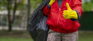 Garbage collector in a red coat and yellow gloves giving a thumbs-up while holding a black trash bag, representing Melbourne Cheap's reliable waste collection services.